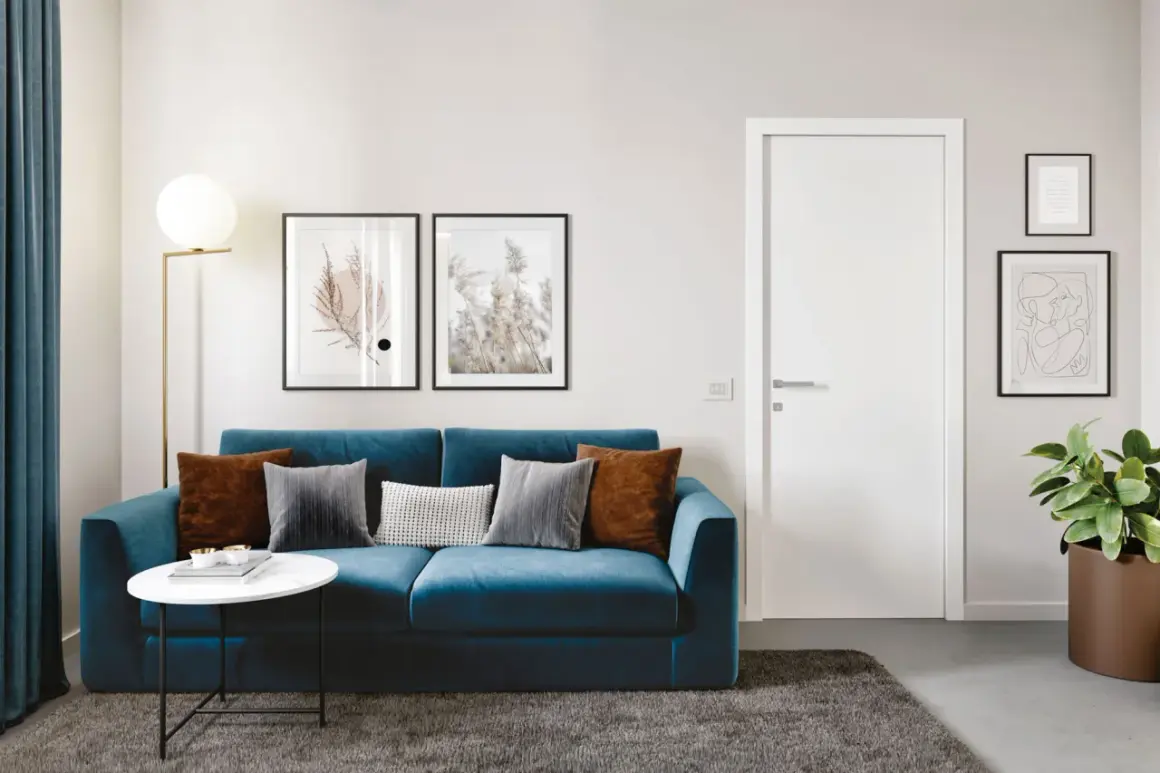 Divano blu acceso con cuscini in velluto tortora e grigio, su un tappeto scuro. Lampada dorata e quadri astratti alle pareti.